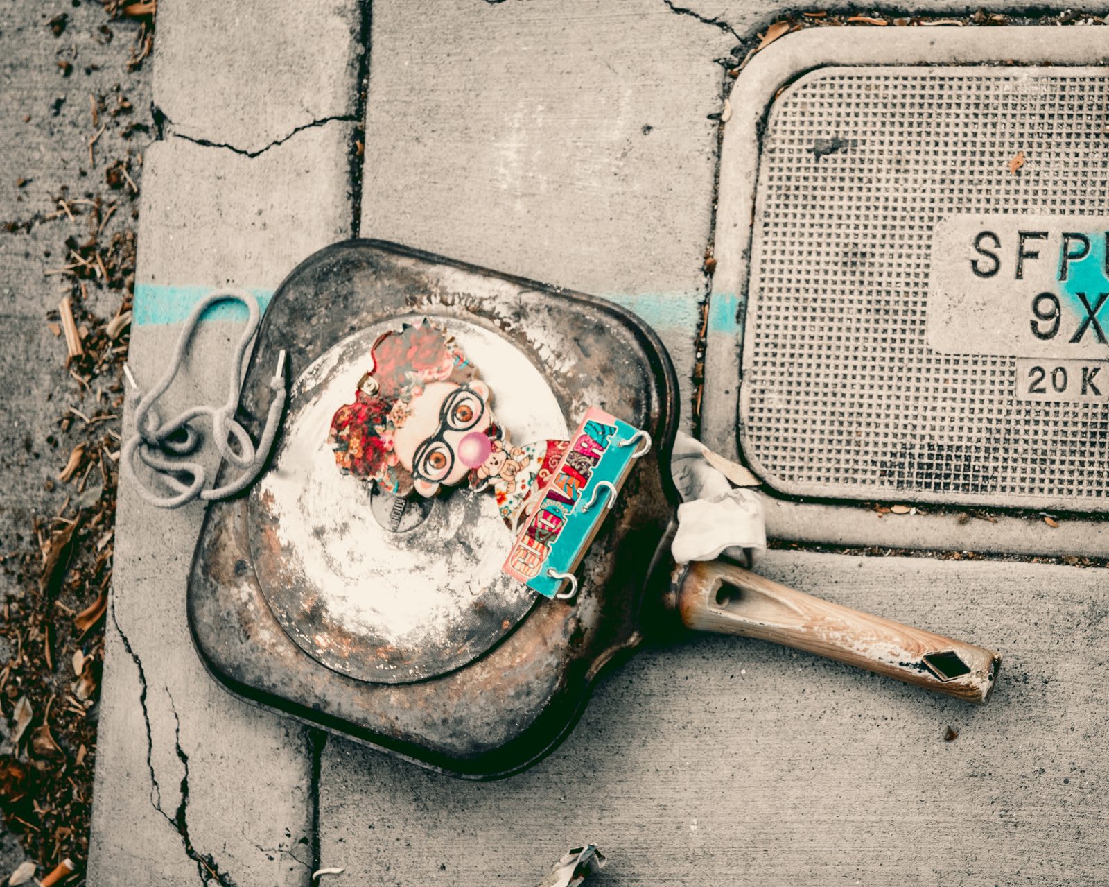 A rusted cast-iron pan abandoned on a cracked sidewalk next to an SFPU sewer cover, a small doll figure resting inside the pan.