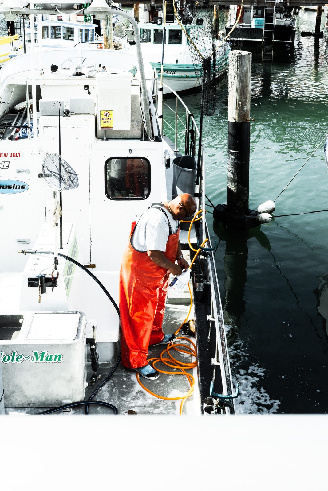 A fisherman in bright orange waders works on his boat at the dock, the green water and other moored boats surrounding him.