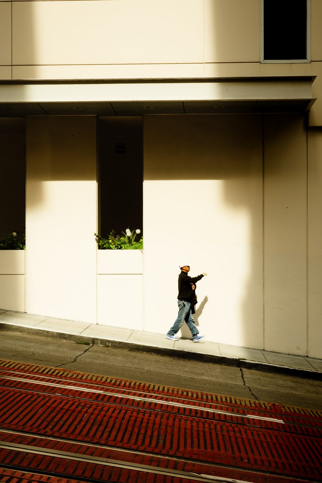 A lone walker in a cap and dark hoodie crossing a long sun-warmed sidewalk above brick cable-car tracks.