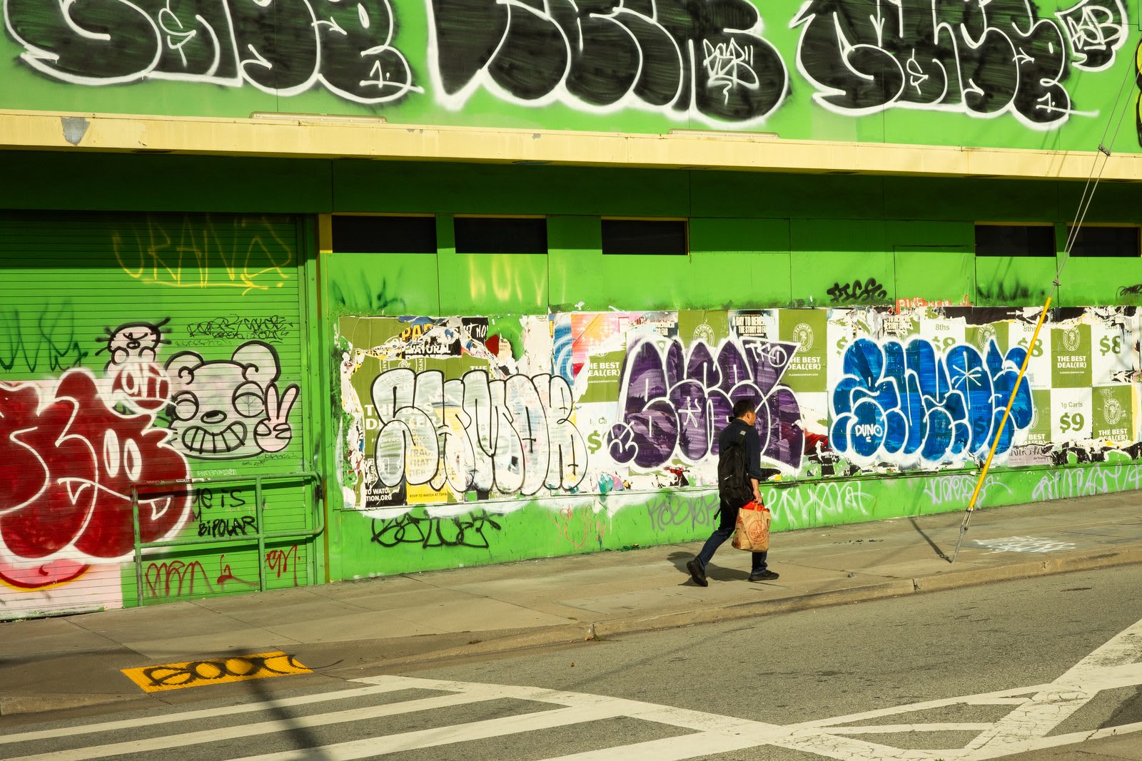 A man walks past a long green wall covered in throw-up graffiti and paste-ups, a row of $9 stickers across the lower half.