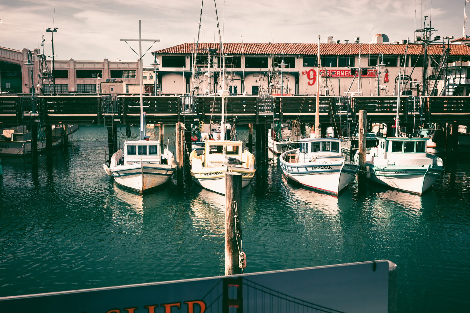 Fishing boats moored in a row at the wharf, their masts and rigging repeating against a pink building with a number 9.