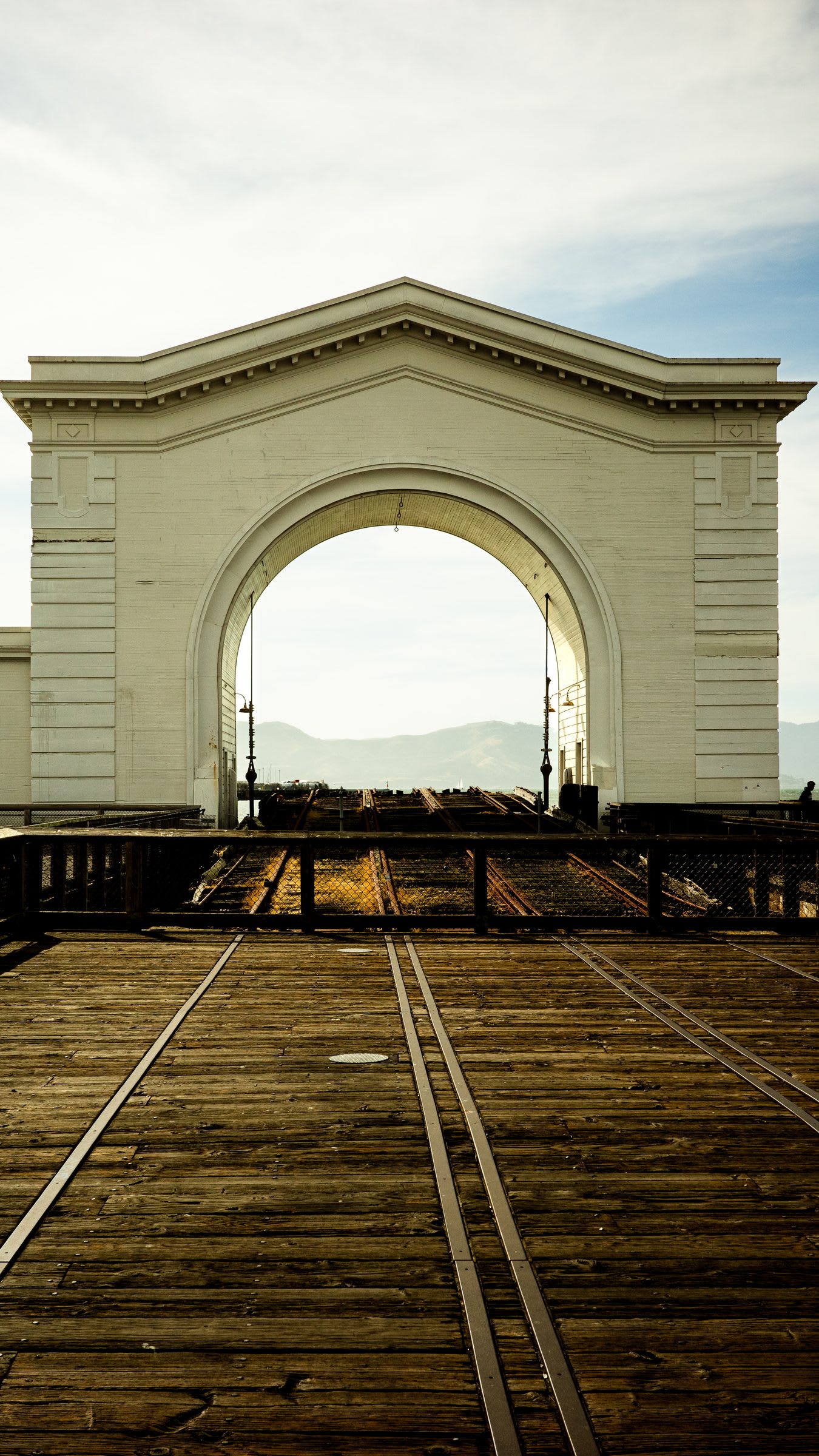 The white Pier 43 ferry arch at the end of a wooden pier, the old rail tracks vanishing through it toward the bay and distant hills.