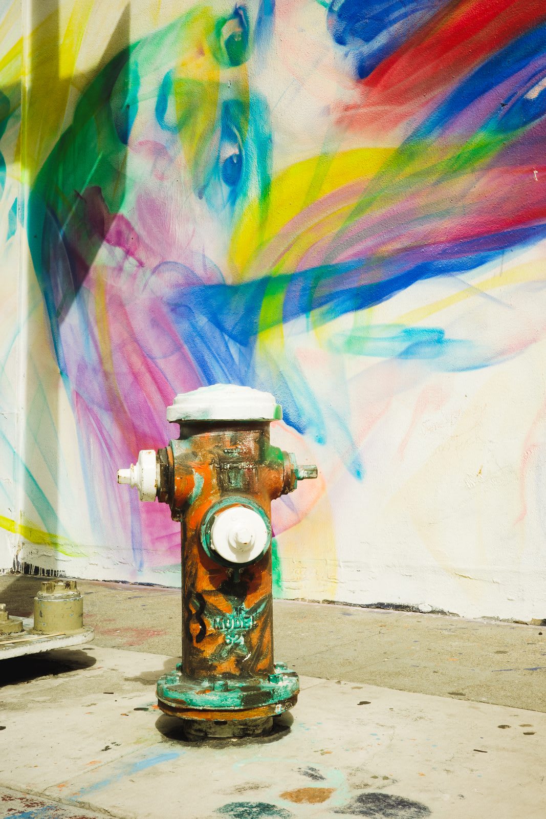 A weathered orange-and-green fire hydrant standing against a wall washed in a rainbow swirl of pastel paint.