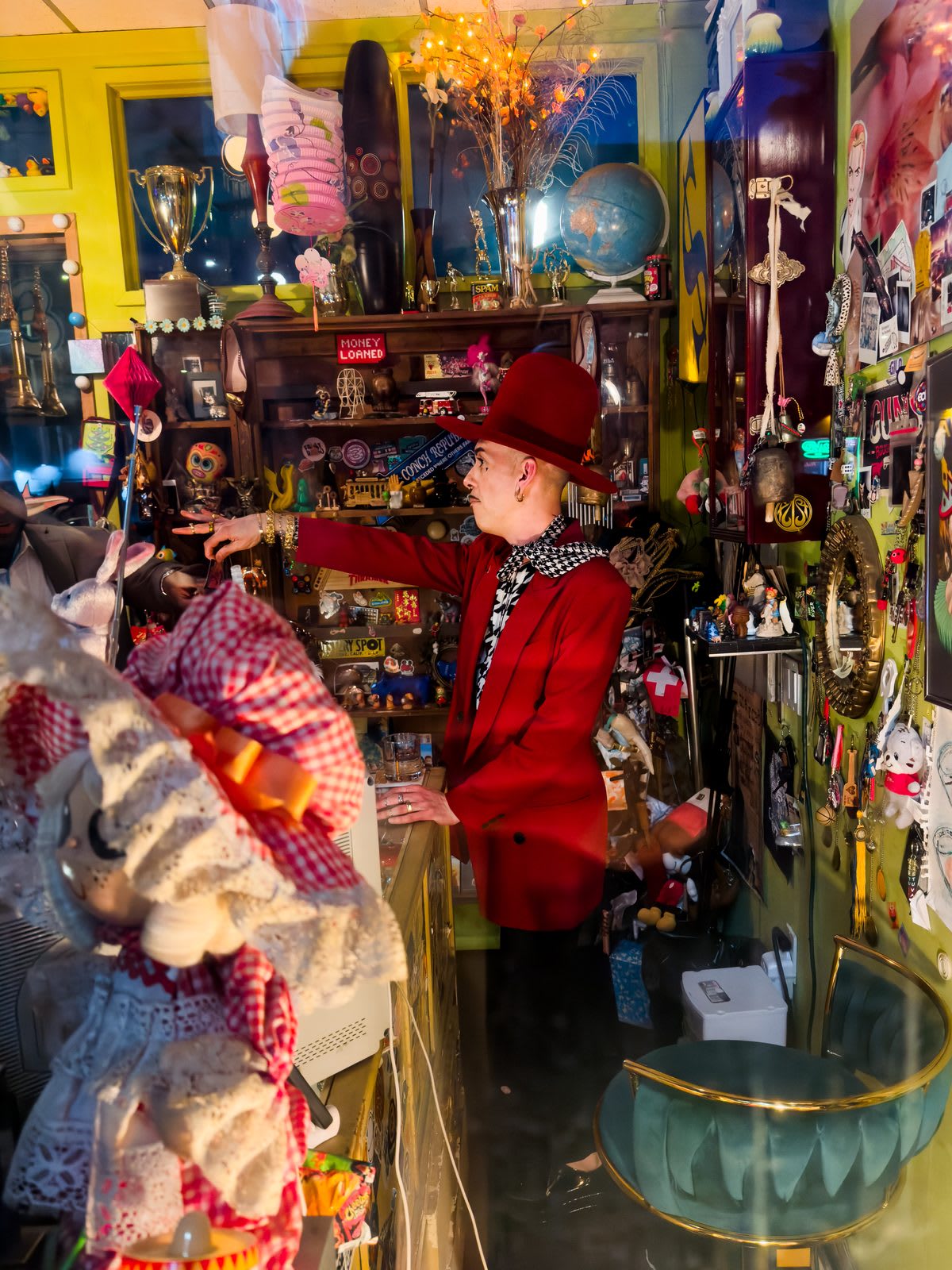 Inside a maximalist vintage curio shop: a shopkeeper in a red felt hat and matching jacket reaches across a counter full of plush dolls and oddities, warm yellow walls behind him.
