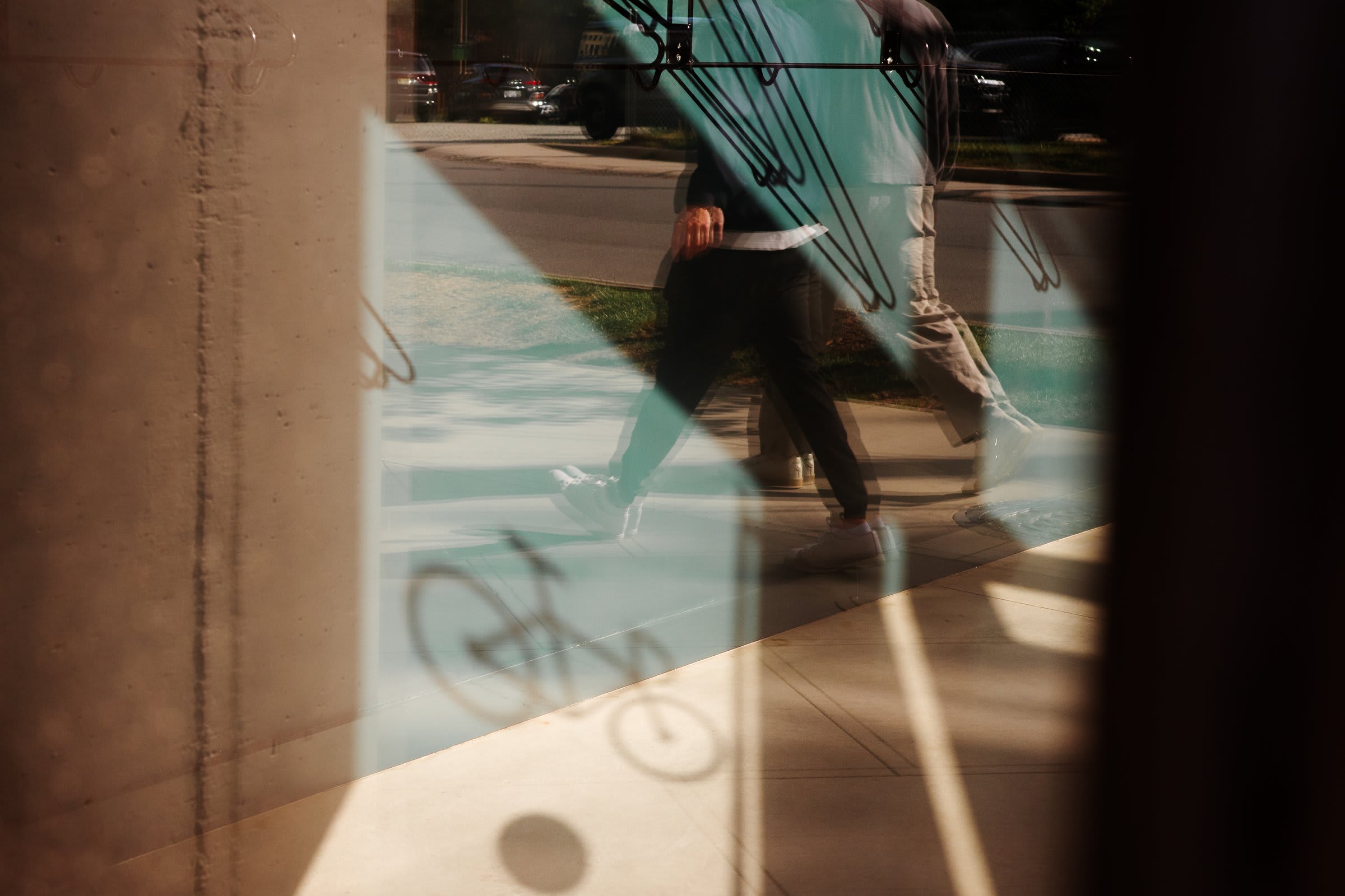 Through a storefront window: a layered reflection of two figures walking past, the silhouette of a bike rack, the street and parked cars beyond.