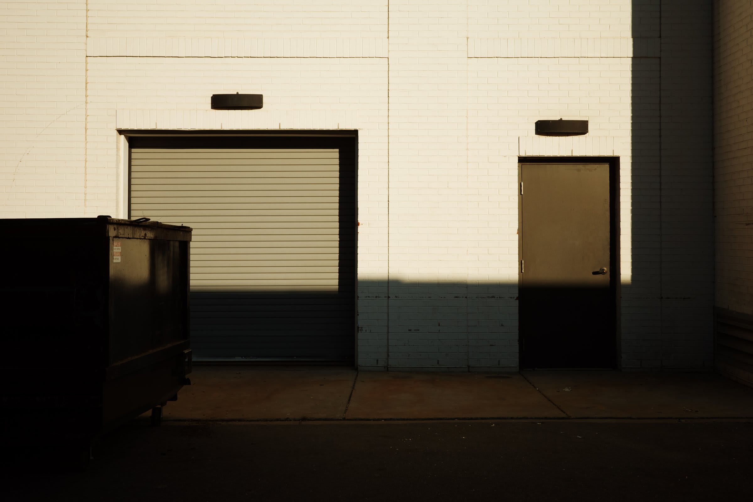 The back of a white-brick warehouse: a roll-up garage door, a steel service door, a dark dumpster, and a hard golden-hour shadow line cutting across the wall.