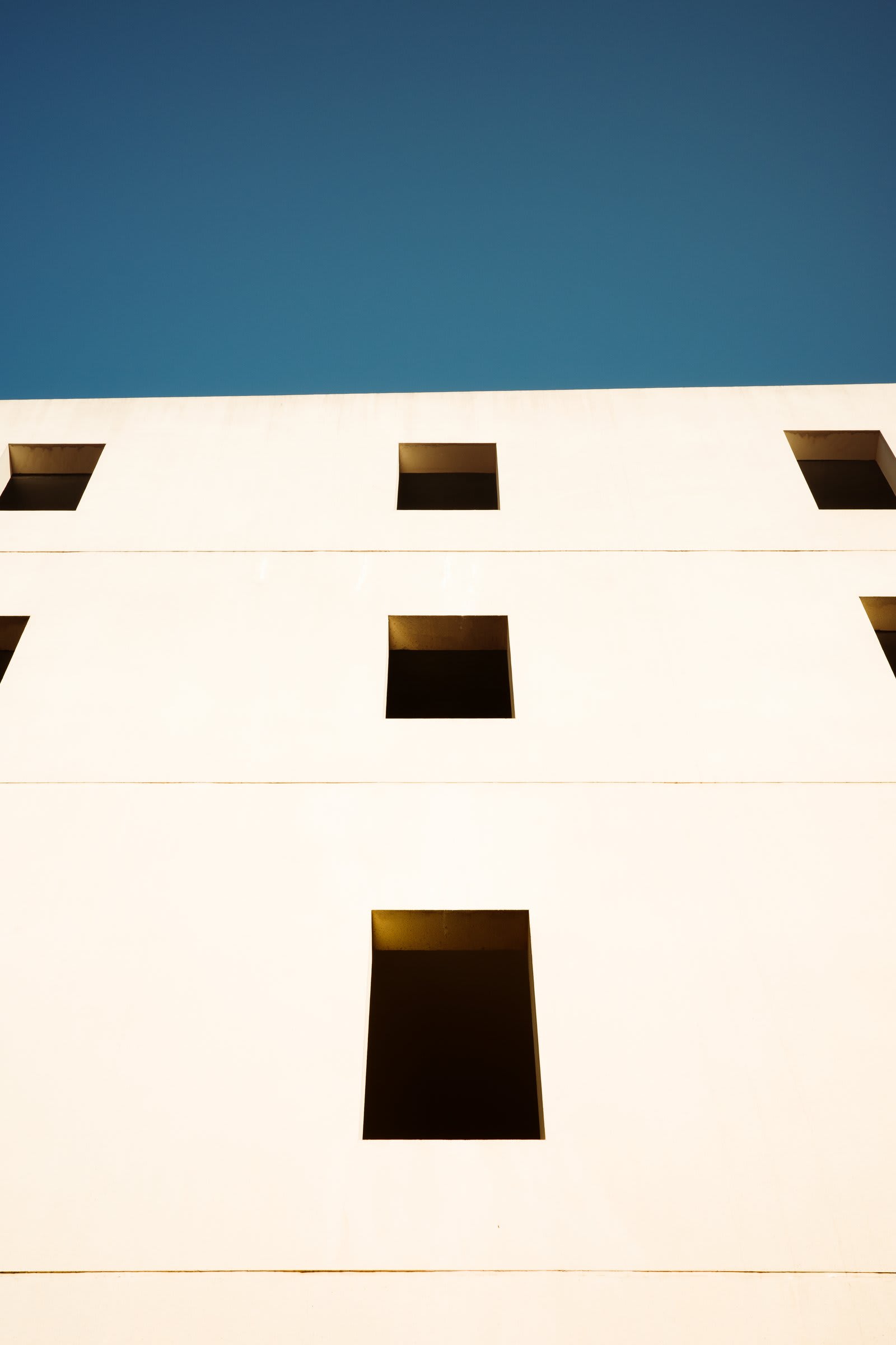 A blank white modernist facade against a deep blue sky, with five dark window cutouts arranged in a sparse grid.