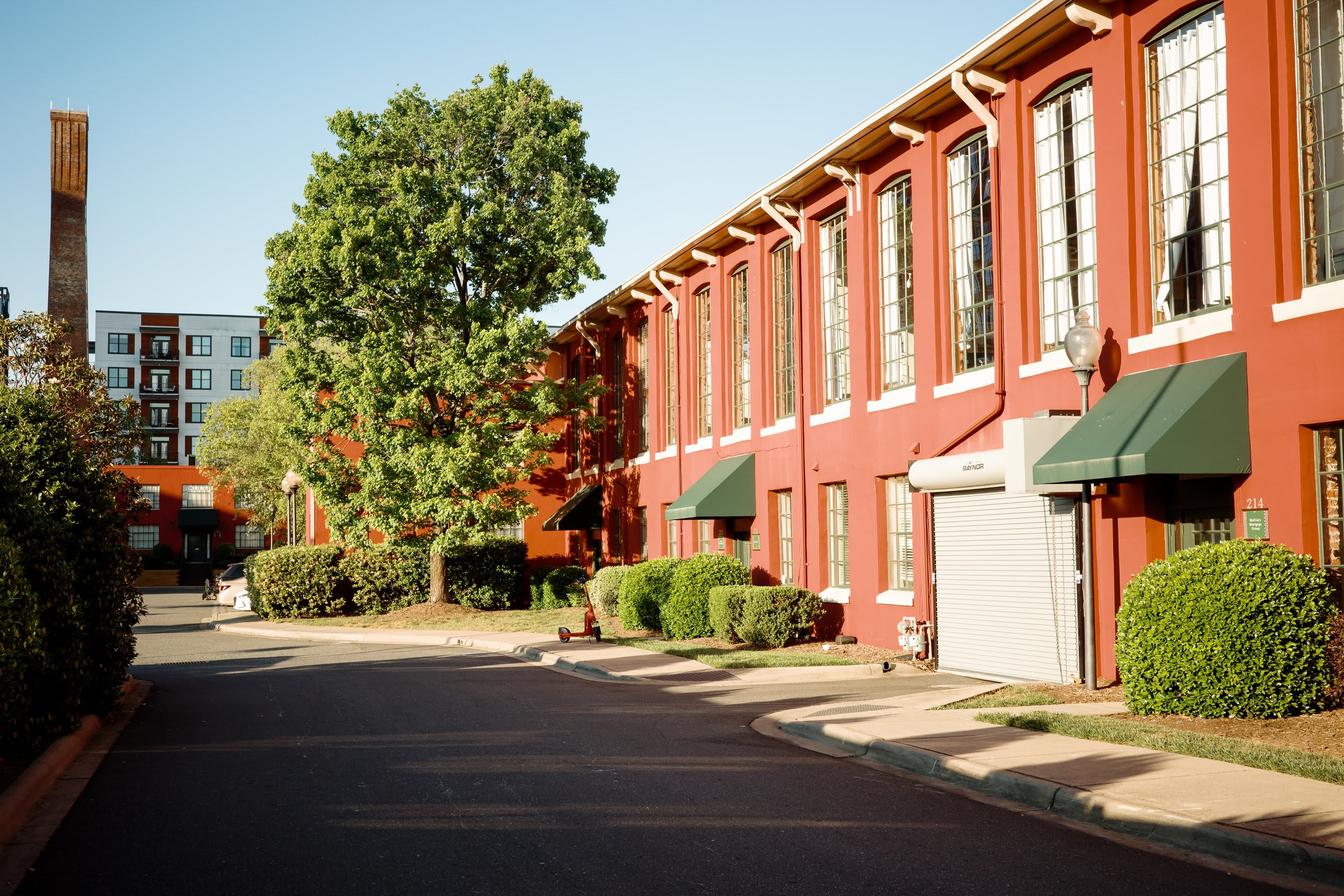 A wide afternoon street view of converted red-brick mill buildings — green awnings, a big tree, and a distant smokestack against a warm sky.