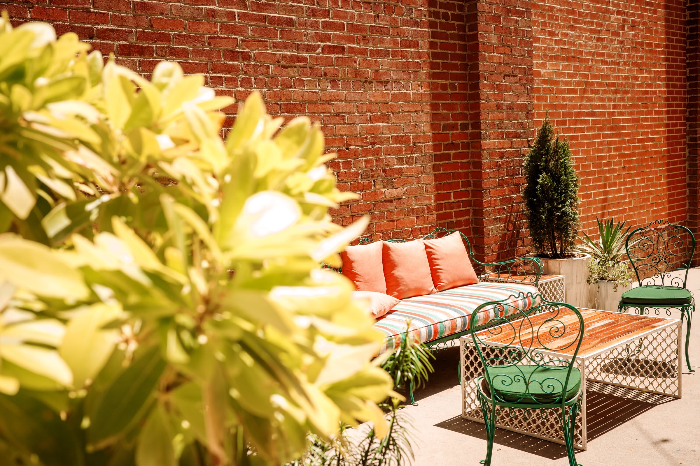 An empty courtyard patio with red brick walls, two green wrought-iron café chairs, a striped cushion bench, and afternoon light pouring in from the left.