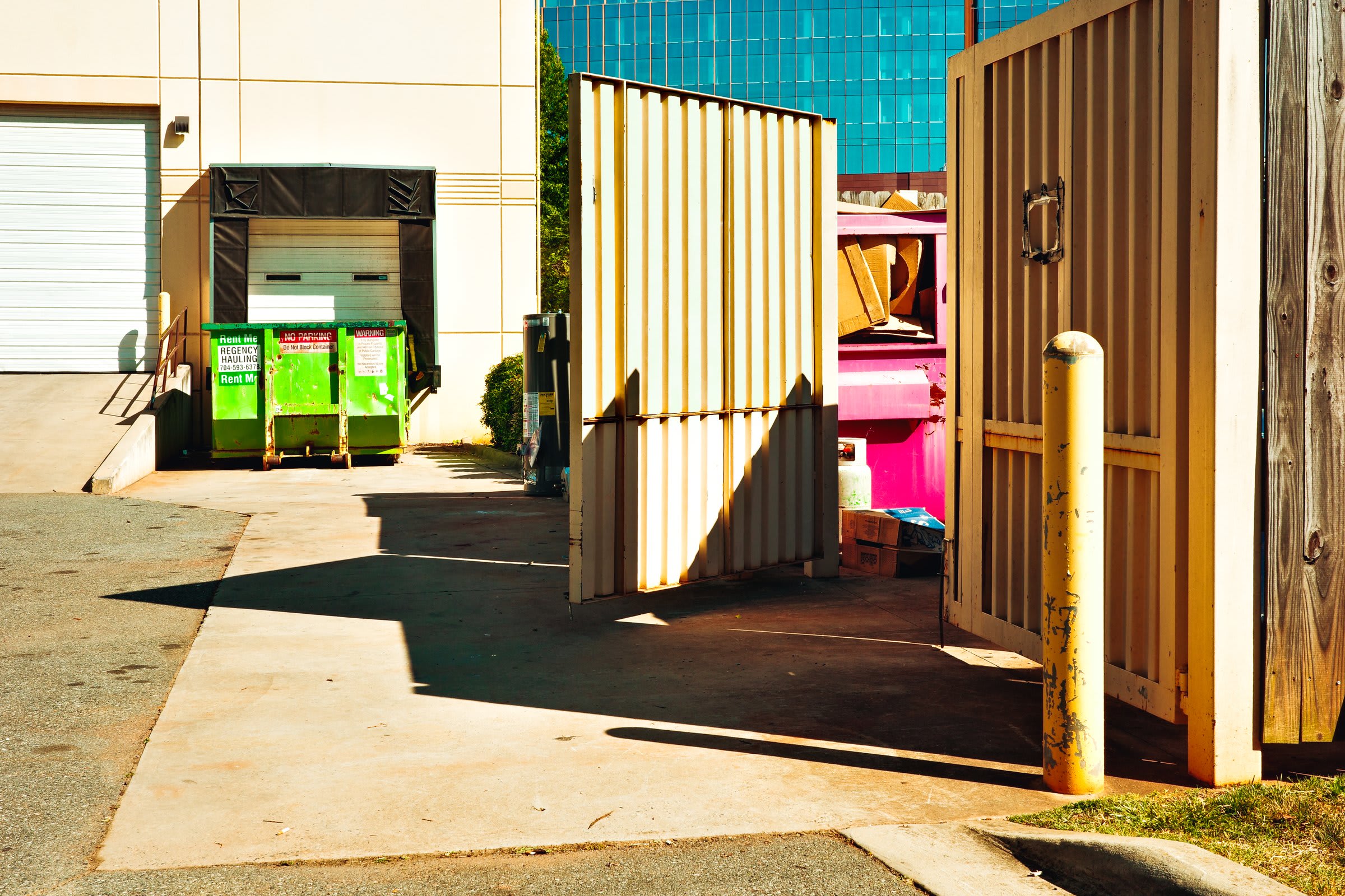 A sunlit alley loading dock: small green and red dumpsters, a hot-pink dumpster peeking past a yellow-bollarded fence, hard mid-afternoon light.