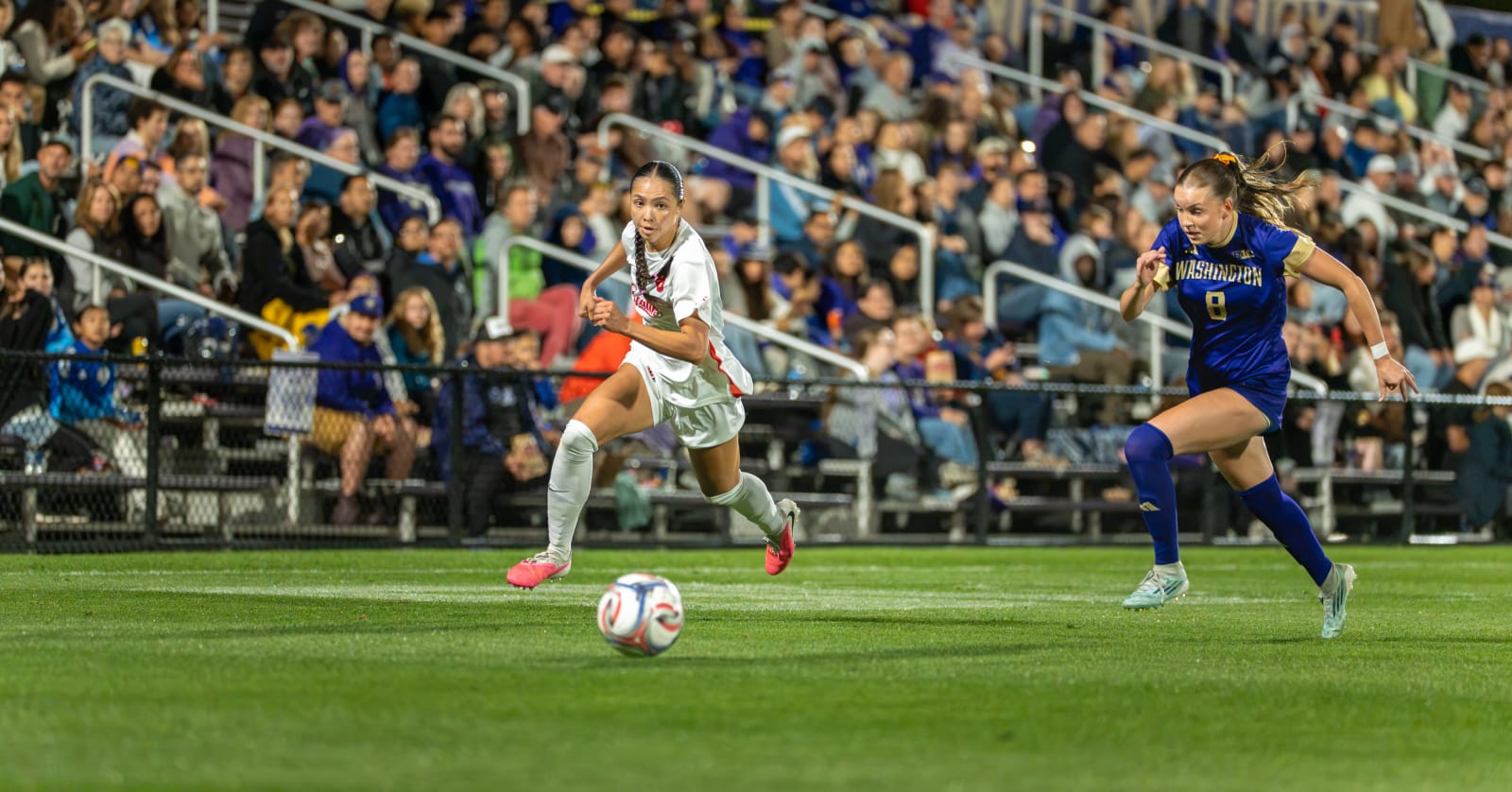 Two college soccer players sprinting for a loose ball under stadium lights, Seattle University women's soccer