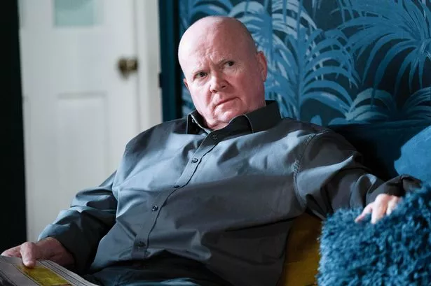 Phil Mitchell reclined in a chair, wearing a blue long-sleeved shirt and looking pensive
