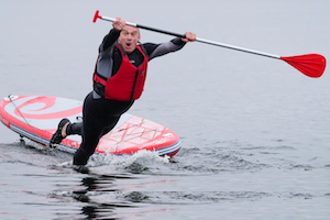 Ed Davey falling off a stand up paddle board and into the water, wearing a wet suit and life jacket and holding a paddle. he looks surprised