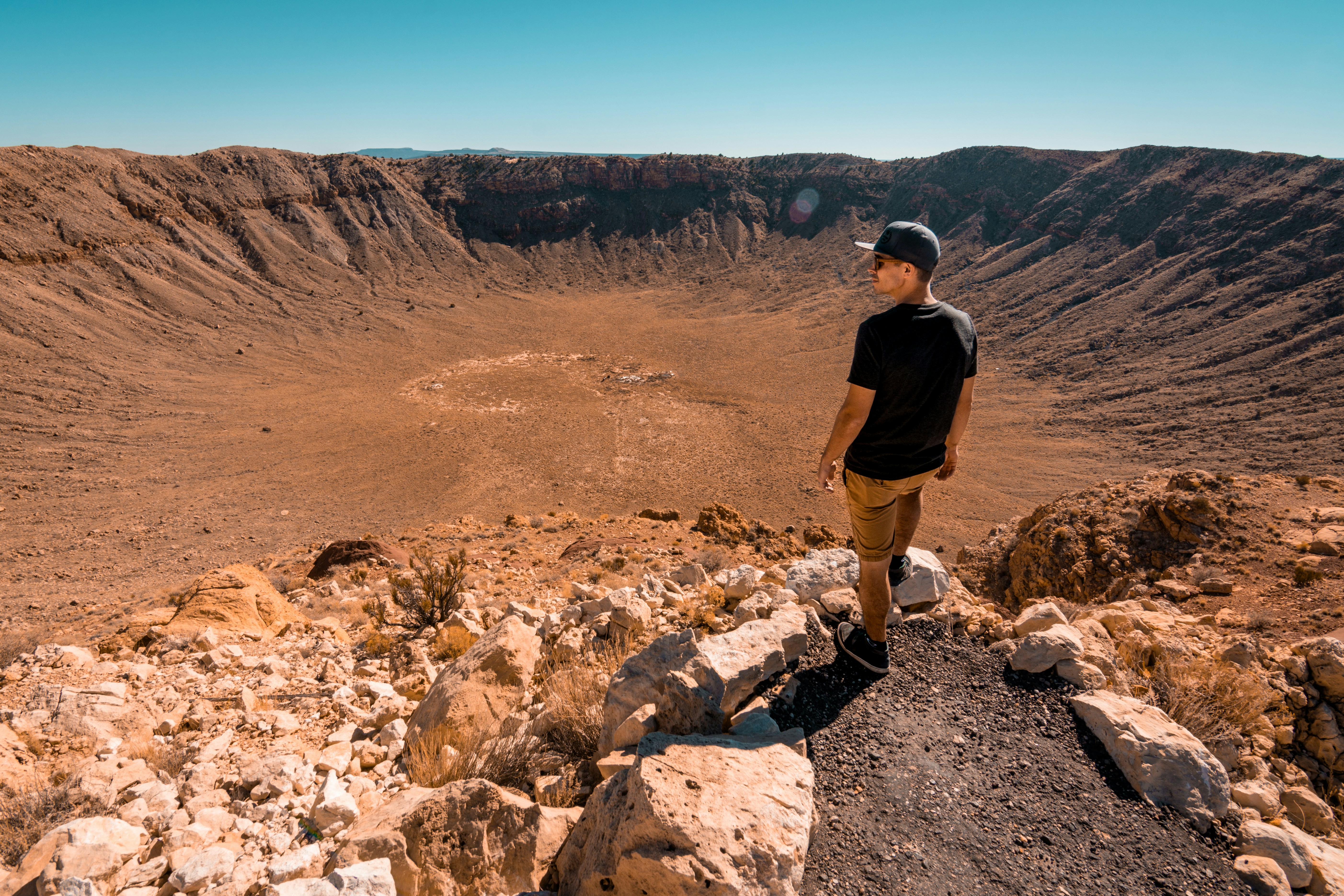 Man wearing shorts, t shirt and cap stands on the edge of huge crater, with bright blue sky in the background