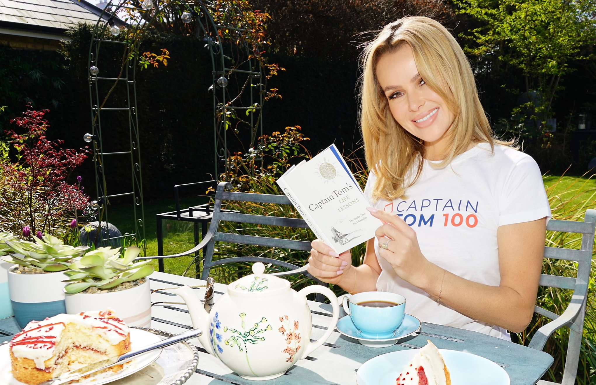 Amanda Holden smiling, reading Captain Tom's book outside at a table in a garden, wearing a white Captain Tom t-shirt
