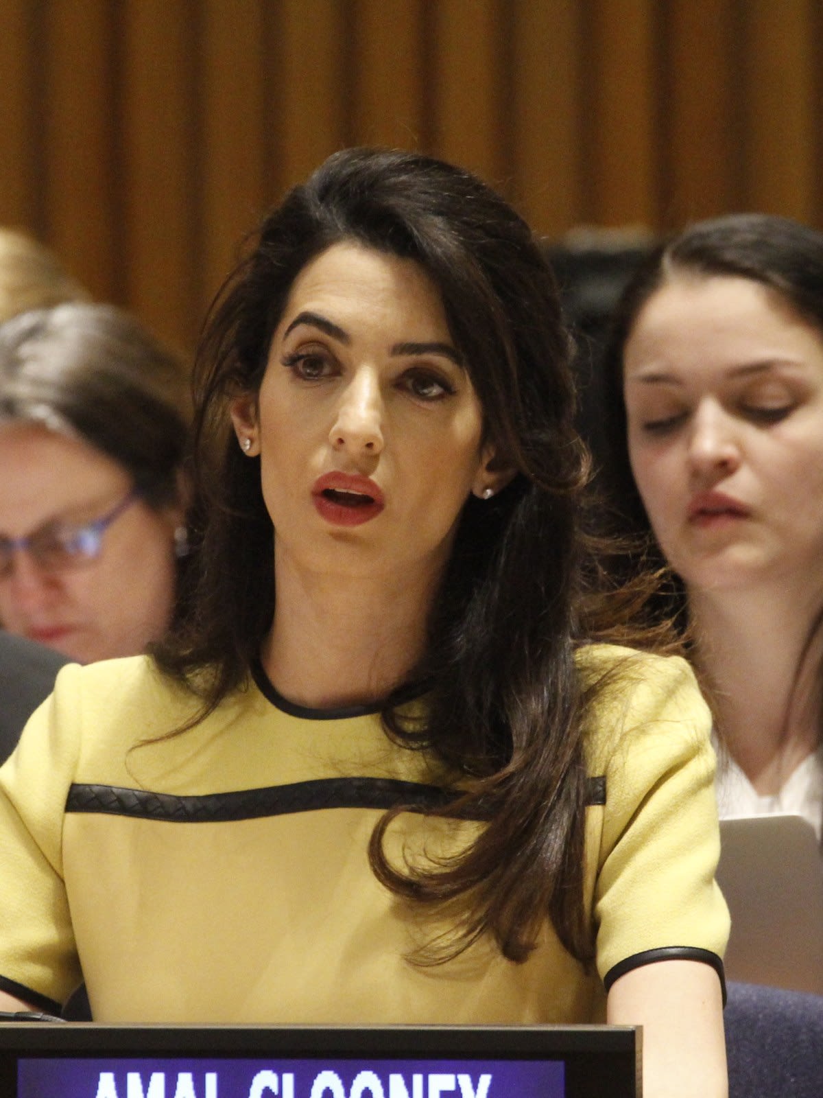 Amal Clooney with long dark hair, wearing a yellow top, seated behind a name plate at the United Nations in 2014