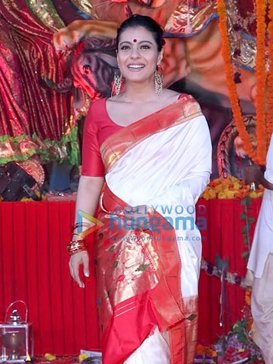 Kajol in a white and red Bengali saree with a red bindi, standing at a Durga Puja pandal in 2018