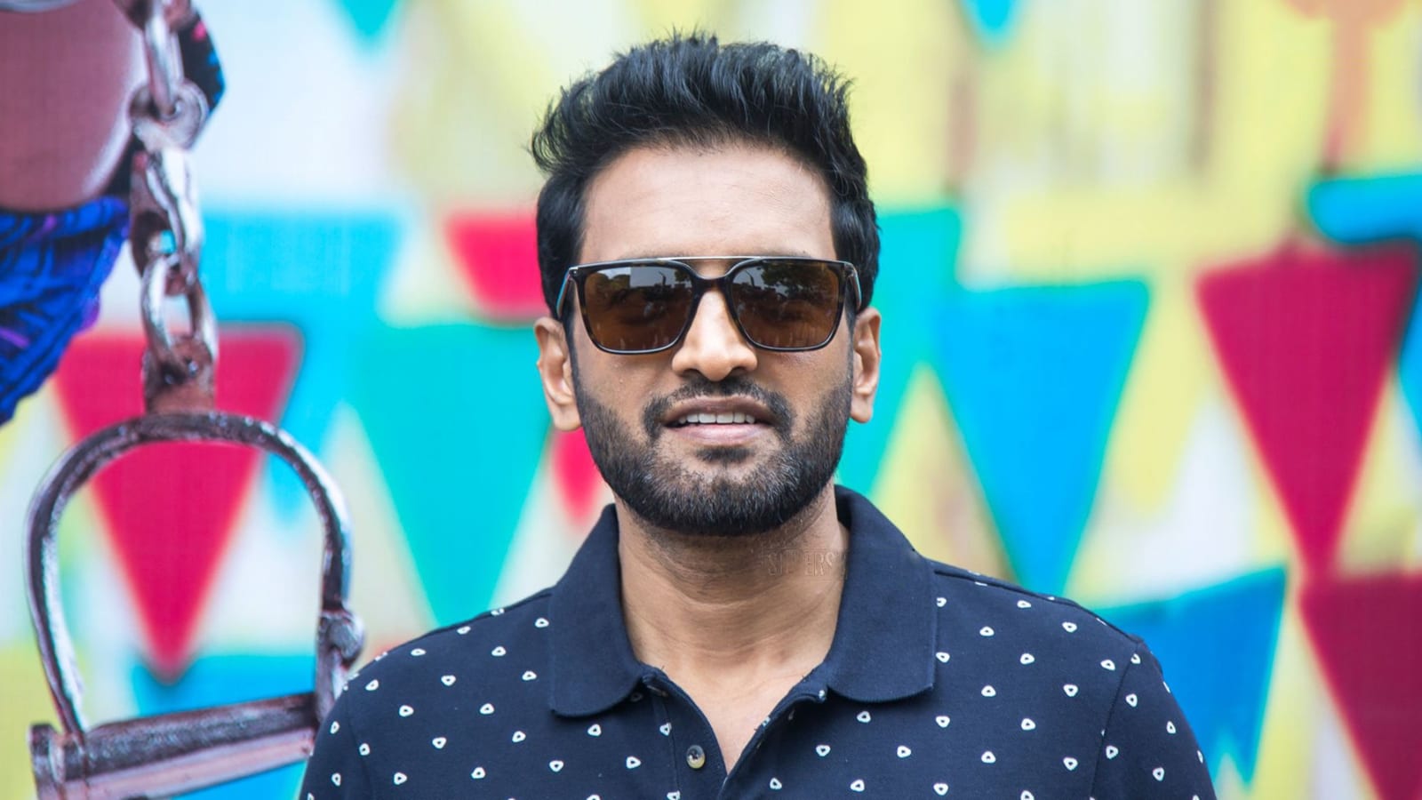 Santhanam in dark sunglasses and a navy patterned polo shirt, smiling in front of a colourful painted backdrop at the A1 press meet in 2019