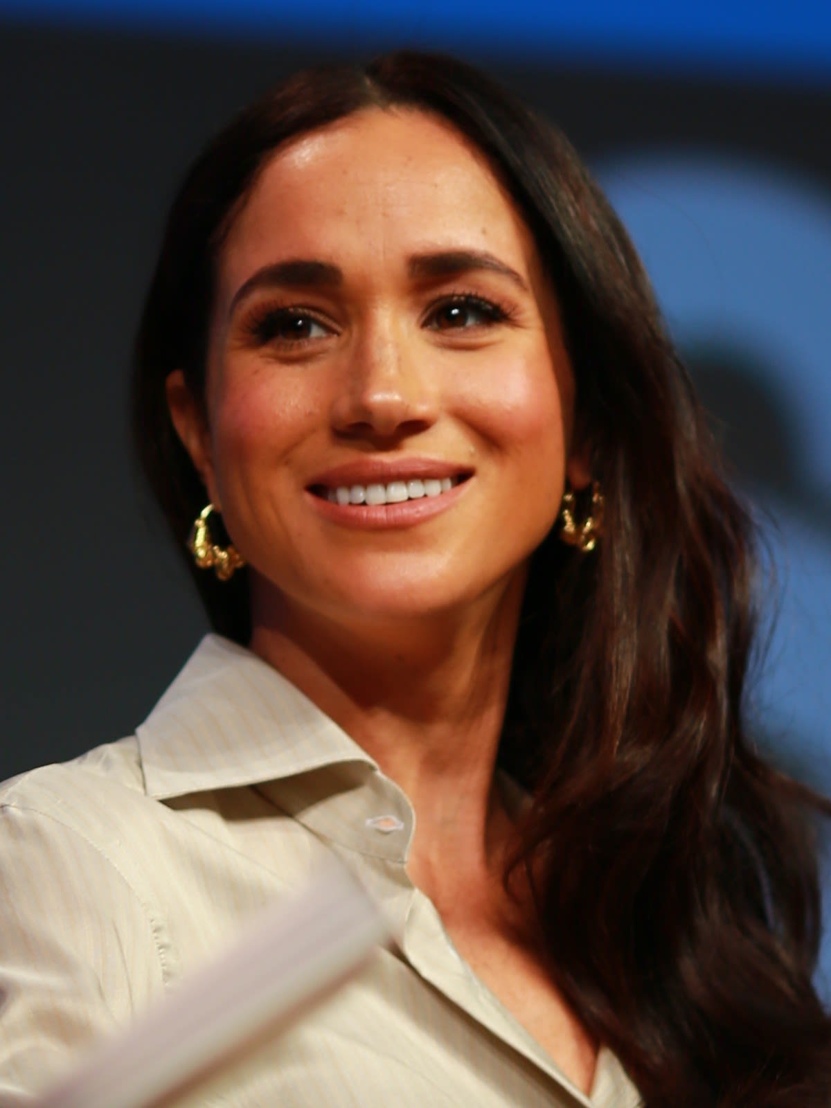 Meghan, Duchess of Sussex, smiling with long dark wavy hair and gold hoop earrings, wearing a cream shirt at SXSW in Austin in 2024