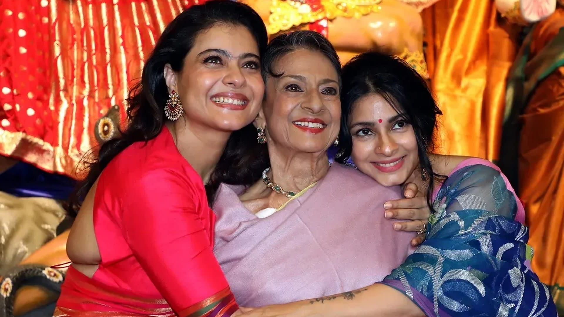 Kajol in a red-orange silk saree and red blouse hugs her mother Tanuja and sister Tanishaa Mukerji in front of the Durga idol at the North Bombay Sarbojanin Durga Puja, Mumbai, October 2026