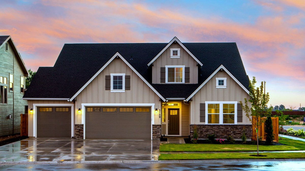 Photorealistic editorial dusk photo of a modern suburban house with warm interior lights, wet driveway reflections and a peach-lavender sky — no people.