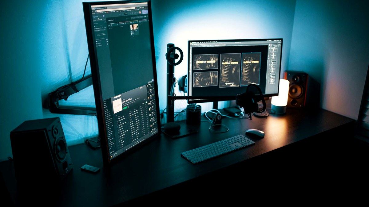 Photorealistic editorial night photo of a dual-monitor developer desk with teal backlight and warm desk lamp, keyboard in foreground, no logos.