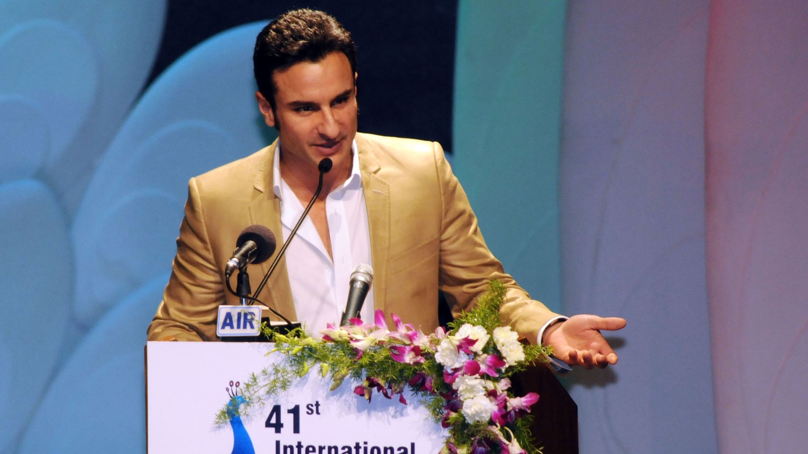 Actor Saif Ali Khan in a gold blazer speaking at a podium at IFFI