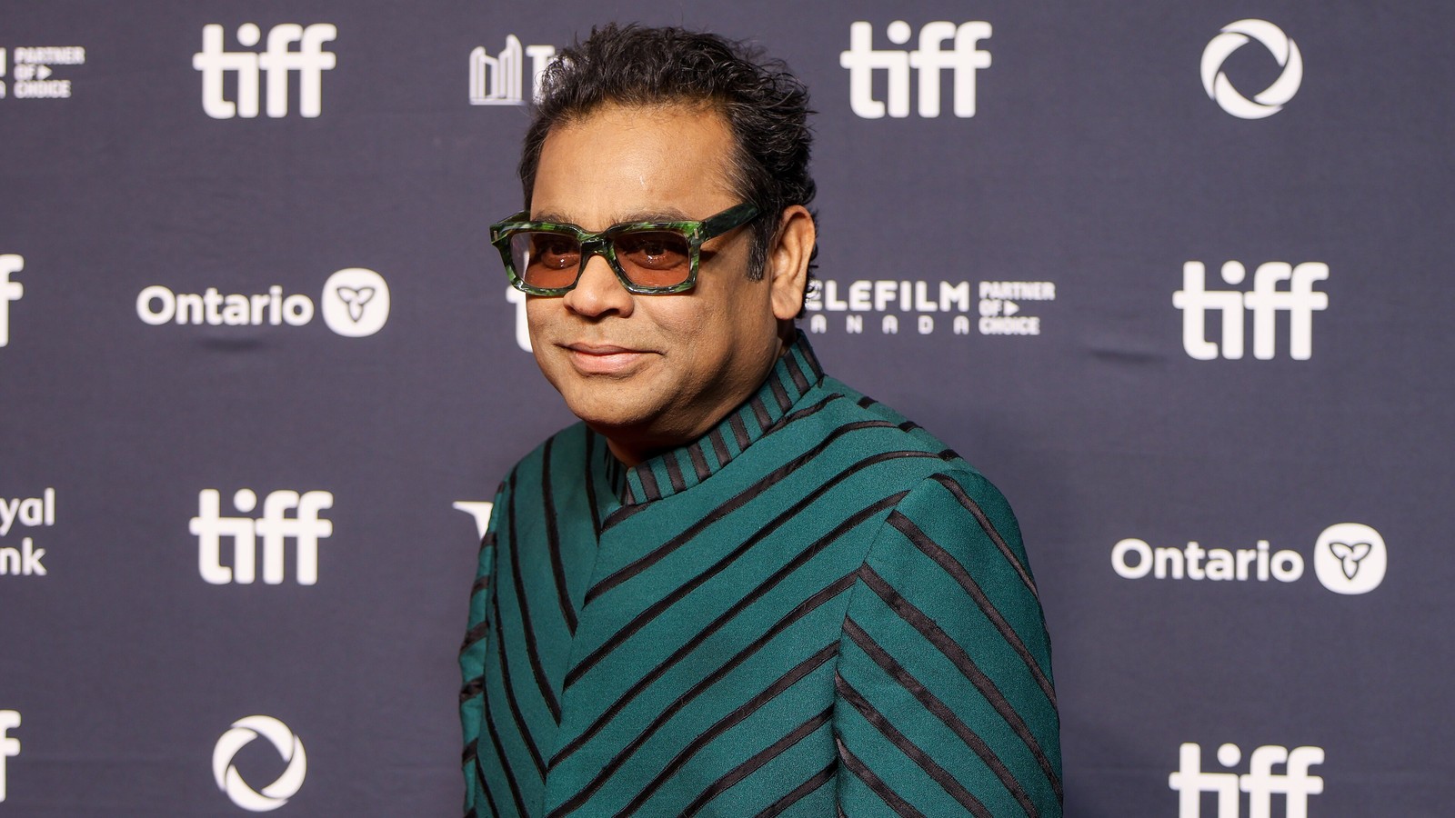 Composer AR Rahman in tinted glasses and a green striped jacket at a TIFF backdrop