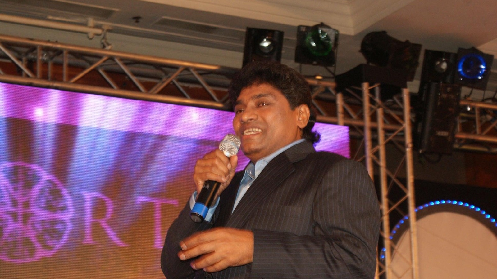 Comedian Johnny Lever holding a microphone on stage in a grey suit