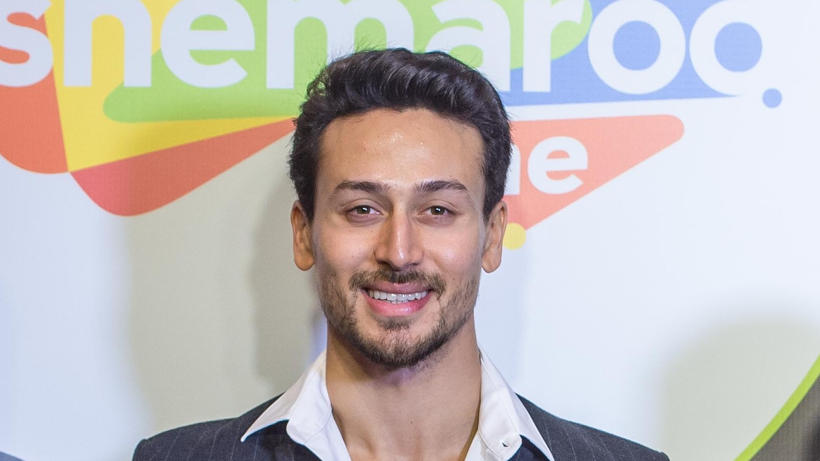Actor Tiger Shroff smiling in a white shirt and pinstriped blazer in front of a ShemarooMe backdrop