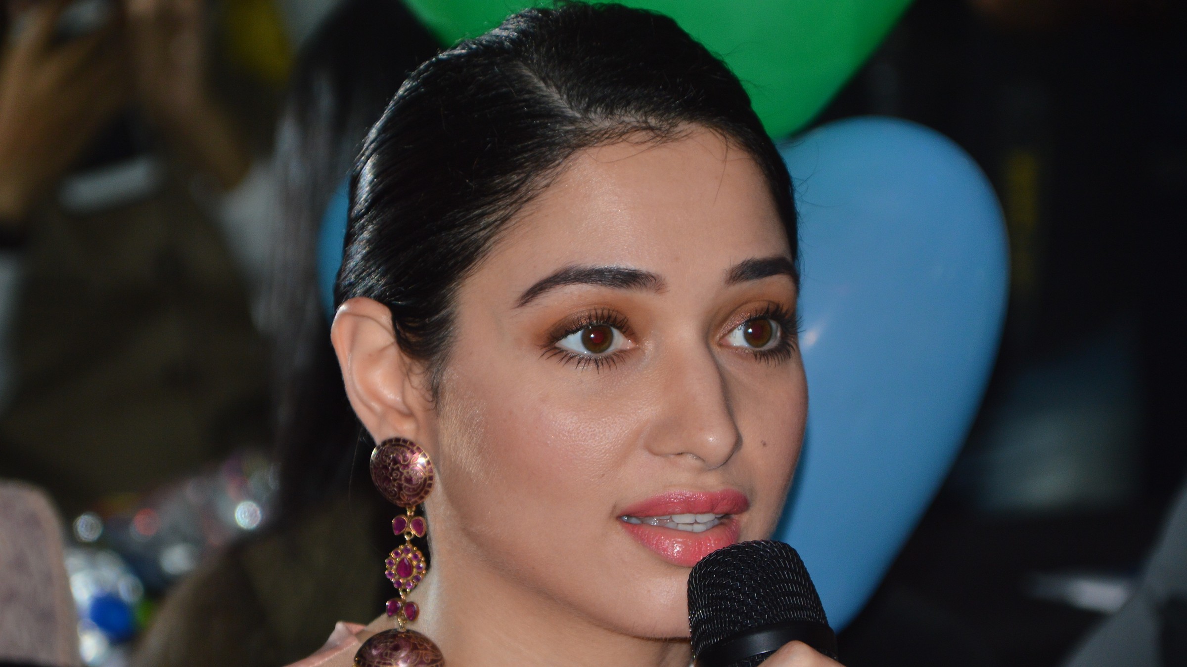 Actress Tamannaah Bhatia with bronze eye makeup and coral lipstick speaking into a microphone at a film event in 2017