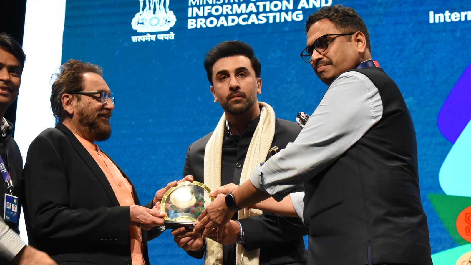Ranbir Kapoor in a dark suit and cream stole receiving a memento on stage at the 55th IFFI in Goa in 2024