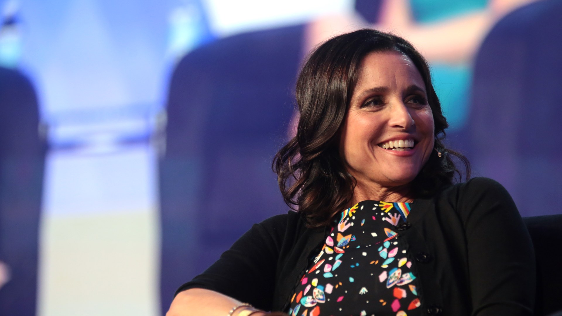 Julia Louis-Dreyfus smiling while seated on stage in a black cardigan and patterned dress at the 2017 WorkHuman conference in Phoenix, Arizona
