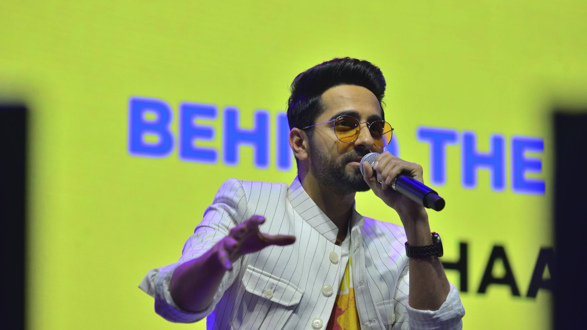 Ayushmann Khurrana in a white pinstriped jacket and tinted round glasses singing into a microphone on stage at IFP Season 8 in 2018