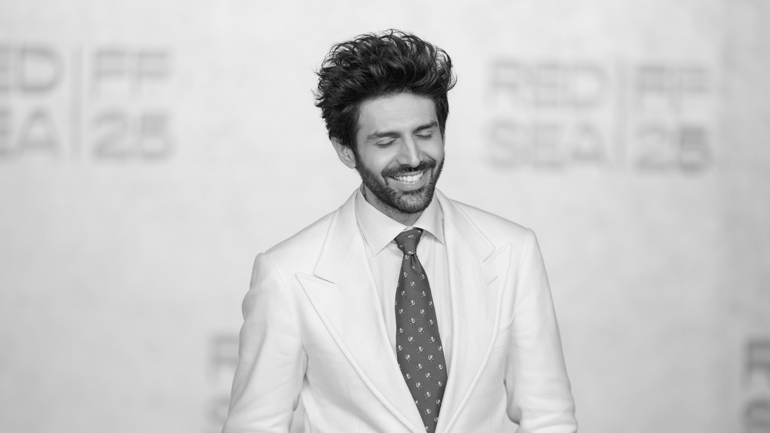Black and white photo of Kartik Aaryan smiling with eyes closed in a white suit and dark patterned tie at the Red Sea International Film Festival 2025
