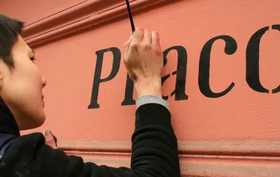 Marta Sieczkowska malująca szyld nad witryną pracowni krawieckiej Teresy Urbanek we Wrocławiu. Marta Sieczkowska painting a sign over Teresa Urbanek Tailor’s Workshop in Wrocław.