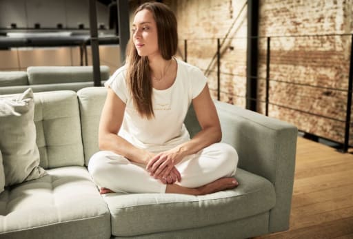 Person in weißer Kleidung sitzt im Schneidersitz auf grauem Sofa in industriell eingerichtetem Raum.