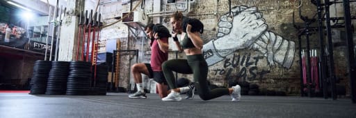 Eine Frau und ein Mann tragen Gewichte auf dem Ruecken und machen Ausfallschritte.