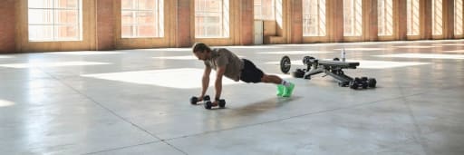 Person macht Liegestütze mit Hanteln in leerer Halle mit Trainingsgeräten im Hintergrund.