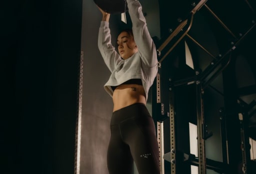 Eine Frau haelt einen Medizinball über Ihren Kopf in Puma Hyrox Trainingskleidung.