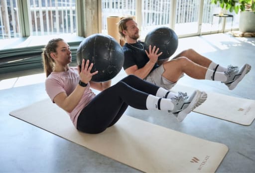 Zwei Personen machen V-Sit-Übungen mit Medizinbällen auf Yogamatten in einem hellen Innenraum mit großen Fenstern.