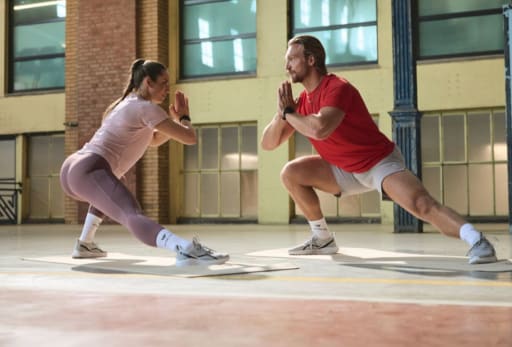 Zwei Personen machen gemeinsam eine Dehnübung im Ausfallschritt in einem Fitnessstudio mit großen Fenstern und Backsteinwänden.