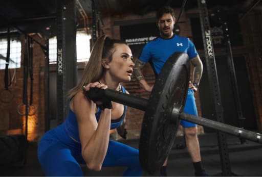  Zwei Personen im Fitnessstudio, eine macht Kniebeugen mit Langhantel, die andere beobachtet.