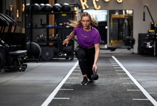 Person im Fitnessstudio macht Kettlebell-Übung im Ausfallschritt, umgeben von Trainingsgeräten.