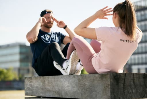 Zwei Personen machen Sit-ups auf einer Holzplattform, Sportoutfit mit ‚WITEBLAZE‘-Logo sichtbar.