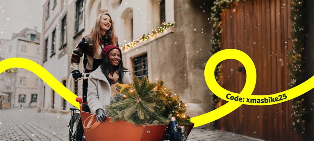 Zwei Frauen fahren mit einem Lastenrad und einem kleinen Weihnachtsbaum durch eine Stadt, in der Schnee fällt.