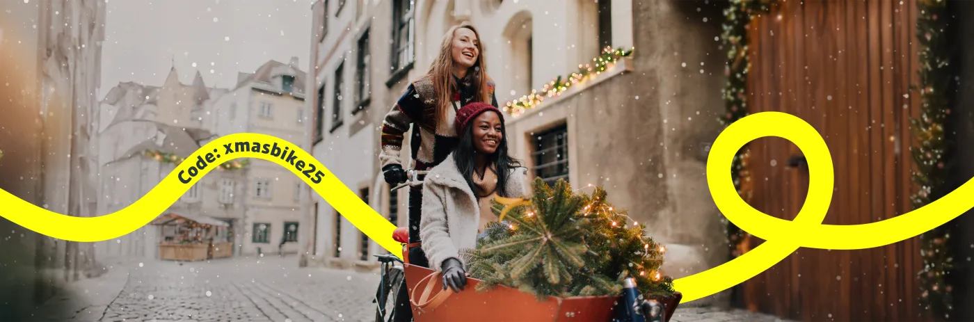 Zwei Frauen fahren mit einem Lastenrad und einem kleinen Weihnachtsbaum durch eine Stadt, in der Schnee fällt.