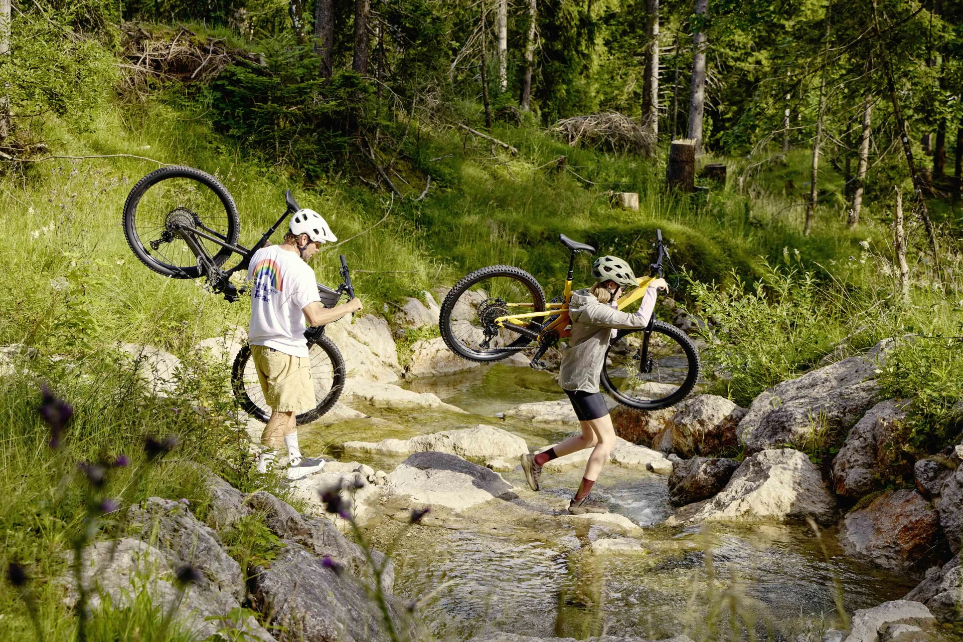 Ein Mann und eine Frau tragen ihre Fully Mountainbikes über einen seichten Flusslauf hinüber.