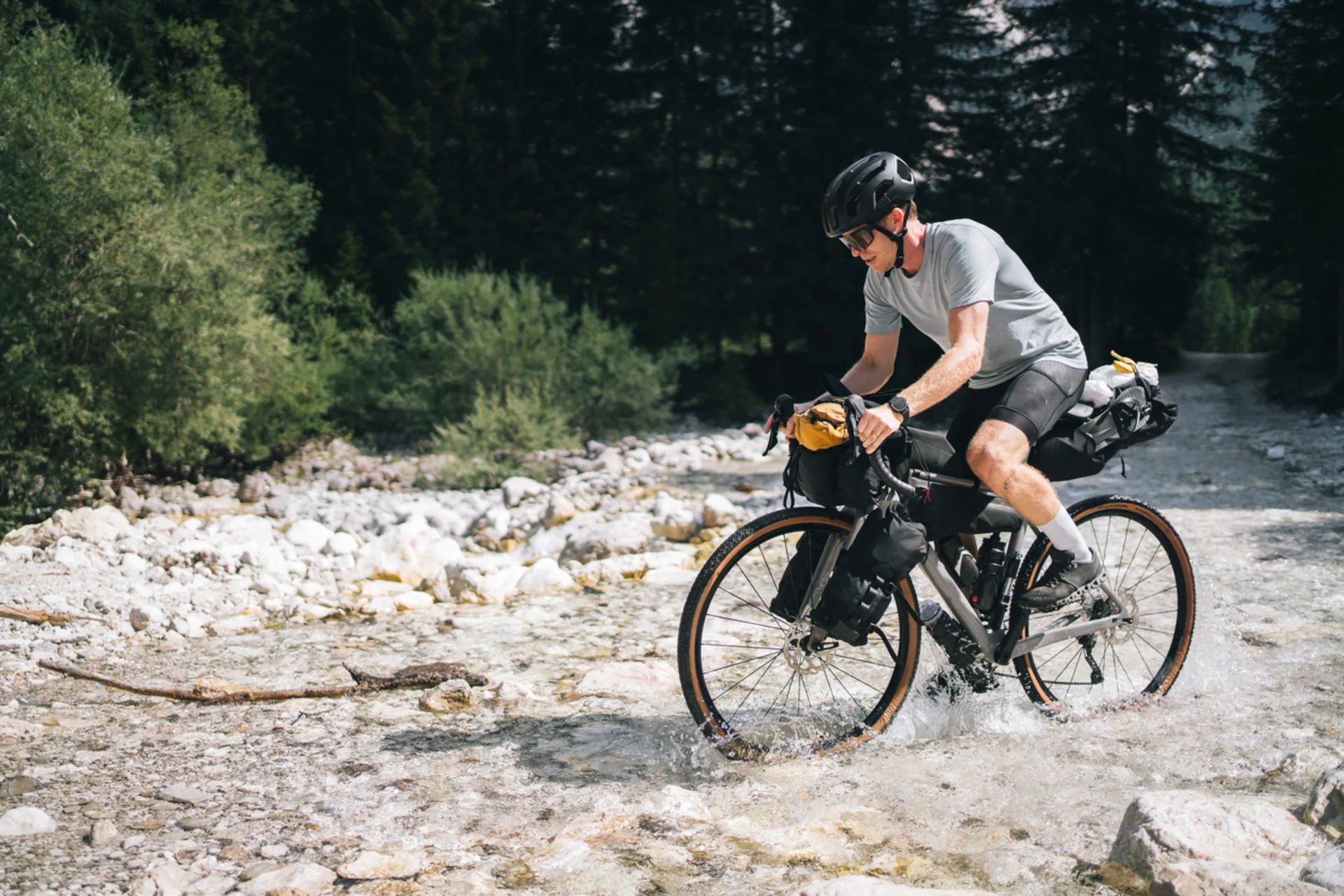 A bike packer wearing a black helmet and riding a fully loaded gravel bike crosses a shallow mountain stream in a rocky landscape.