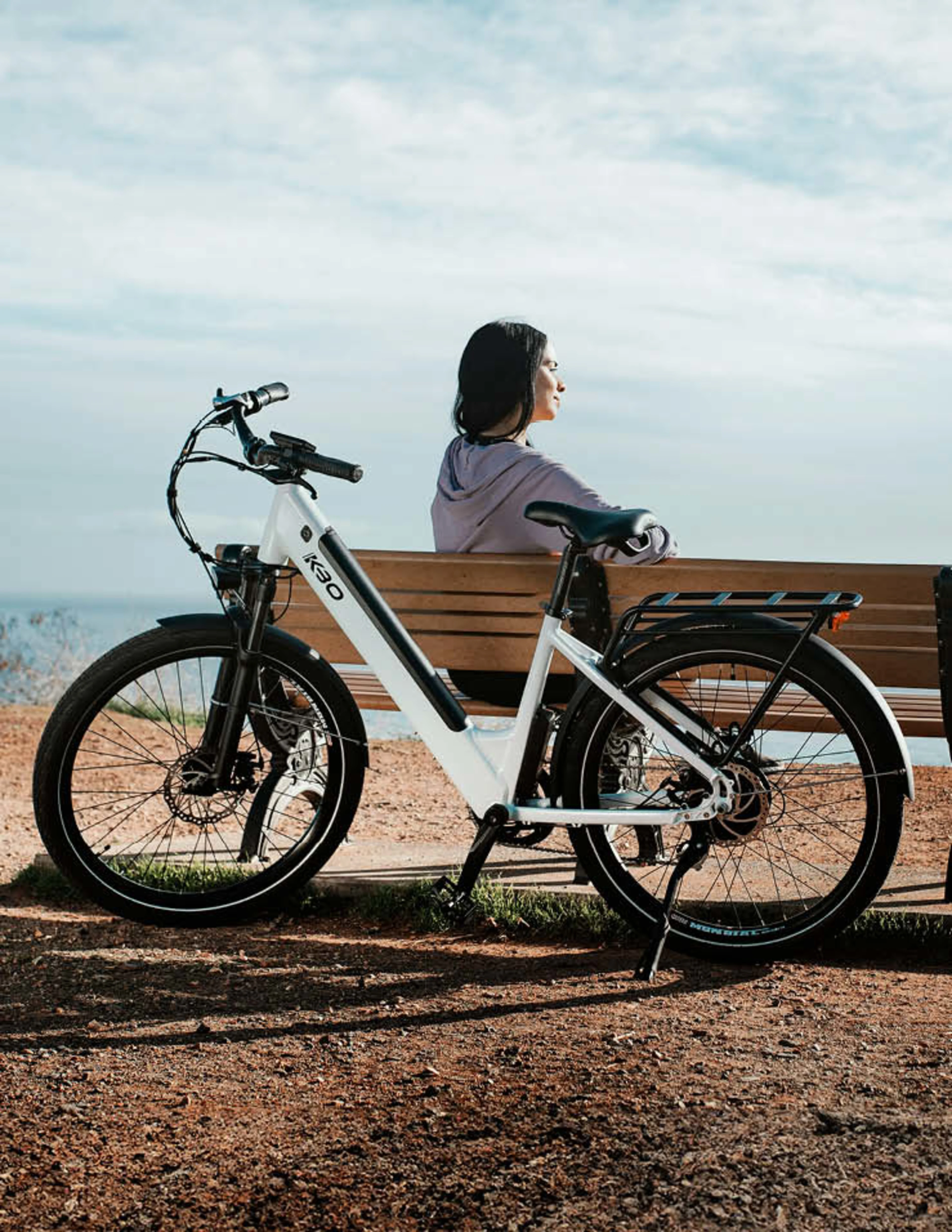 Eine Frau sitzt auf einer Bank auf einem Hügel und schaut in die Ferne. Hinter ihr steht ein E-Bike.